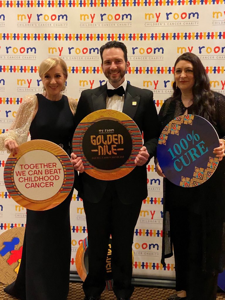 Three people stand in front of a My Room branded media wall. There is a woman on the left, a man in the middle and a woman on the right. They are all in formalwear and are holding signs about beating childhood cancer, and aiming for a cure.