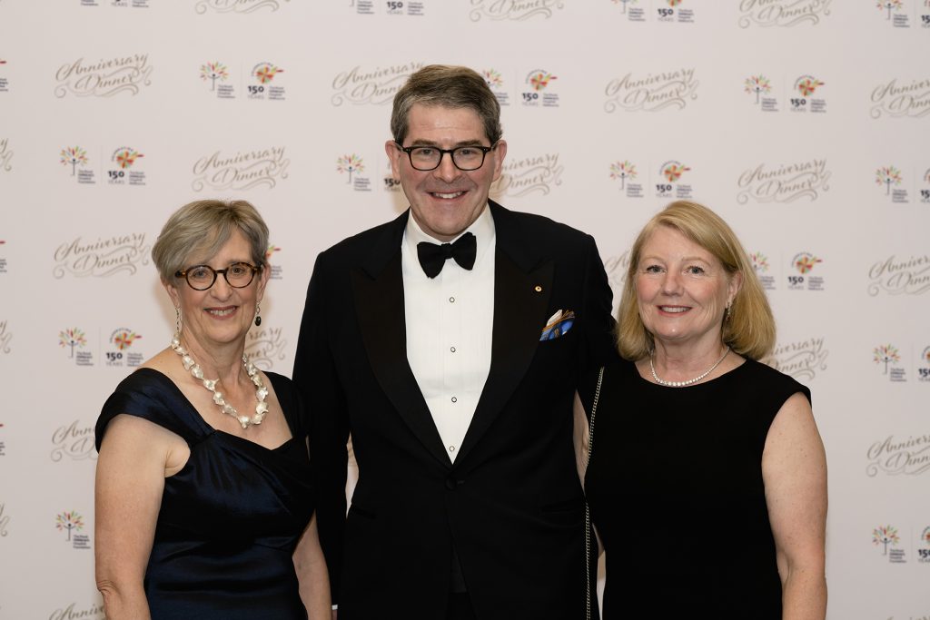 Three people stand in front of a media wall. The wall is branded with Anniversary Dinner and the Royal Children's Hospital Foundation logo. There is a woman on the left, a man in the middle and a woman on the right. They are all smiling and dressed in formalwear. 