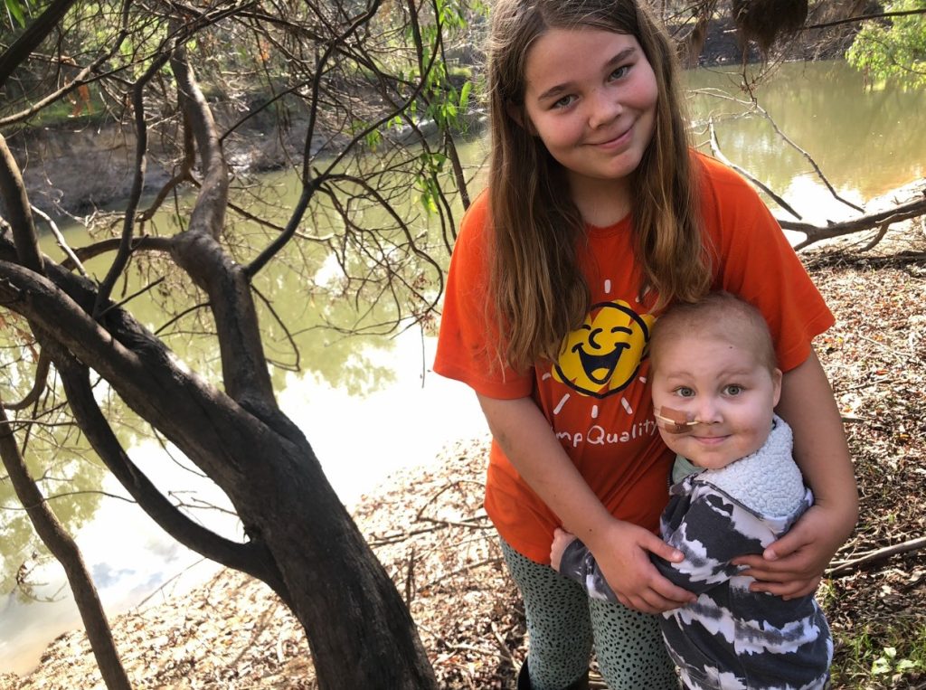A young girl and boy standing in front of a river. The girl is older. She is wearing an orange shirt with Camp Quality on it. She is hugging the boy. He has a shaved head and a nasogastric tube. 